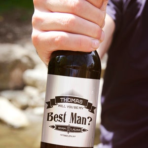 May include: A brown glass bottle with a white label that says "Thomas Will You Be My Best Man? Adam & Laura October 12th, 2017".