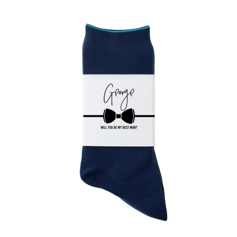 Pu&ograve; includere: Un paio di calzini blu scuro con un'etichetta bianca che dice "George" e "Vuoi essere il mio testimone?" con un papillon nero.