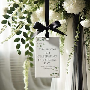 Peut inclure: Une étiquette rectangulaire avec le texte "THANK YOU FOR CELEBRATING OUR SPECIAL DAY!" est suspendue par un ruban noir noué en nœud. L'étiquette présente un motif aquarelle de feuilles vertes et les noms "CELENE AND NOAH". Le fond présente des fleurs blanches et de la verdure.