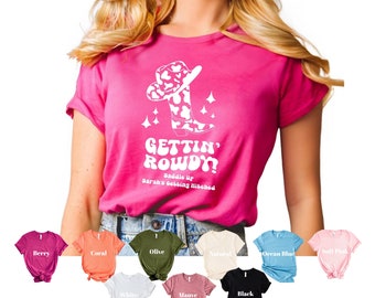Op de afbeelding: Een roze T-shirt met een witte afbeelding van een cowboylaars en een hoed met de tekst "Gettin' Rowdy! Saddle up Sarah's getting hitched". Het shirt is verkrijgbaar in verschillende kleuren, waaronder bessen, koraal, olijf, naturel, oceaanblauw, zacht roze, wit, mauve en zwart.