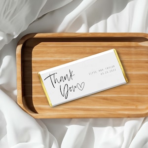 May include: White chocolate bar with gold foil wrapping. The bar has black text that says "Thank You" with a heart symbol and "Elyse and Taylor 05.25.2022".