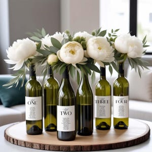 Könnte beinhalten: Sechs grüne Weinflaschen mit weißen Etiketten, angeordnet auf einem Holztablett. Jede Flasche enthält weiße Blumen und Grünzeug. Die Etiketten sind von eins bis vier nummeriert, mit zusätzlichem Text darunter.
