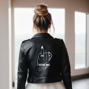 Peut inclure: Une veste en cuir noir avec un motif blanc au dos. Le motif représente une main avec une bague au majeur, faisant un doigt d'honneur, et le texte "FUTURE MRS Smith".