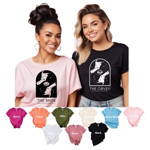 Peut inclure: Deux femmes portant des t-shirts avec un motif graphique de mains et un œil dans une arche. Le texte sur les t-shirts est "The Bride" et "The Coven". Les t-shirts sont disponibles dans une variété de couleurs, y compris le rose, le noir, le corail, l'olive, le naturel, le bleu océan, le rose pâle, le blanc, le mauve et le noir.