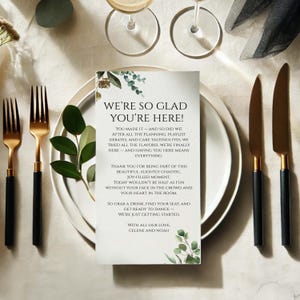 Peut inclure: Mise en place de mariage avec une assiette blanche, des couverts à manche doré et une carte avec le texte "WE'RE SO GLAD YOU'RE HERE!". La carte est ornée de motifs botaniques verts.