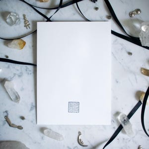 May include: A blank white card with a small, square, gray design in the lower center. The card is surrounded by crystals, black ribbon, and small silver charms on a marble surface.
