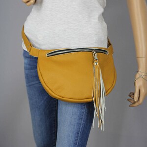 May include: A mustard yellow leather fanny pack with a white fringe tassel. The fanny pack has a black zipper and a silver zipper pull.