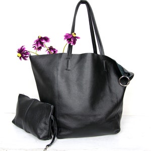 May include: A black leather tote bag with a smaller black leather pouch attached. The tote bag has two handles and a silver ring on the side. The pouch has a zipper closure. The bag is sitting on a white surface with purple flowers.