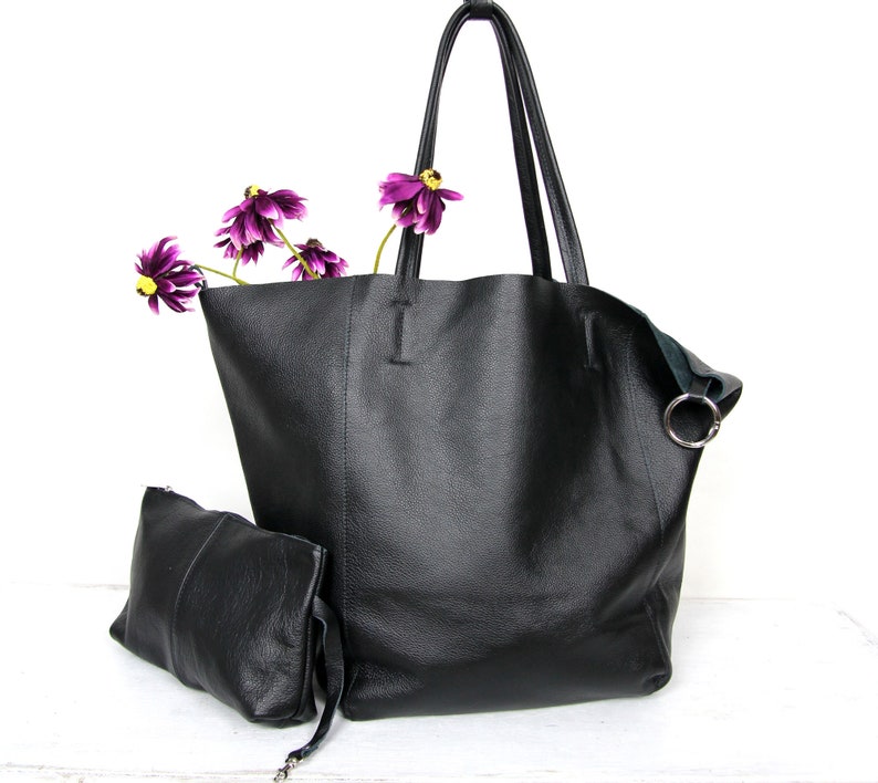 May include: A black leather tote bag with a smaller black leather pouch attached. The tote bag has two handles and a silver ring on the side. The pouch has a zipper closure. The bag is sitting on a white surface with purple flowers.