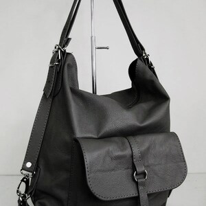 K&ouml;nnte beinhalten: Dunkelgraue Lederhandtasche im Rucksackstil mit einer Vordertasche und verstellbaren Tr&auml;gern. Die Tasche hat ein weiches, l&auml;ssiges Design mit silberfarbenen Beschl&auml;gen. Die Tr&auml;ger k&ouml;nnen als Rucksack oder Umh&auml;ngetasche getragen werden.