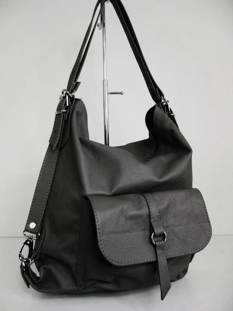 K&ouml;nnte beinhalten: Dunkelgraue Lederhandtasche im Rucksackstil mit einer Vordertasche und verstellbaren Tr&auml;gern. Die Tasche hat ein weiches, l&auml;ssiges Design mit silberfarbenen Beschl&auml;gen. Die Tr&auml;ger k&ouml;nnen als Rucksack oder Umh&auml;ngetasche getragen werden.