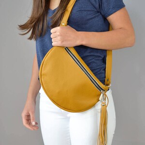 May include: A mustard yellow leather fanny pack with a black zipper and a long white and brown tassel. The bag is worn across the body.