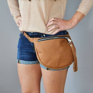 May include: A brown leather fanny pack with a zipper closure and a silver tassel. The fanny pack is worn around the waist of a person wearing denim shorts.