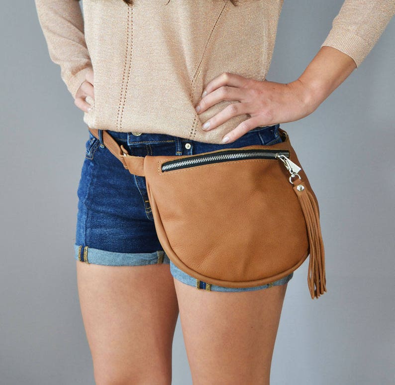 May include: A brown leather fanny pack with a zipper closure and a silver tassel. The fanny pack is worn around the waist of a person wearing denim shorts.