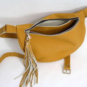 May include: A mustard yellow leather fanny pack with a white zipper and a white tassel. The bag has a buckle closure and a long strap.