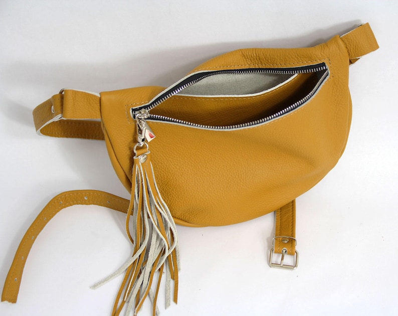 May include: A mustard yellow leather fanny pack with a white zipper and a white tassel. The bag has a buckle closure and a long strap.