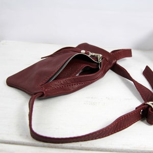 Puede incluir: Un bolso bandolera de cuero burdeos con cierre de cremallera y una larga correa ajustable. El bolso est&aacute; abierto y el interior es visible.