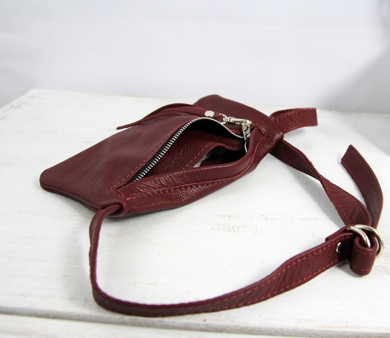 Puede incluir: Un bolso bandolera de cuero burdeos con cierre de cremallera y una larga correa ajustable. El bolso est&aacute; abierto y el interior es visible.