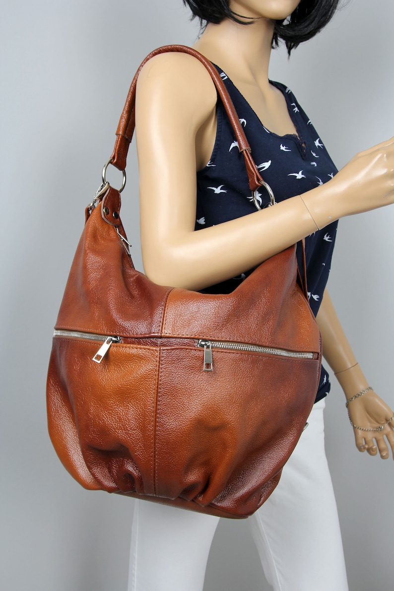 Pu&ograve; includere: Una borsa a tracolla in pelle marrone con una tasca con cerniera e un tiretto della cerniera argentato. La borsa ha un aspetto casual e morbido.