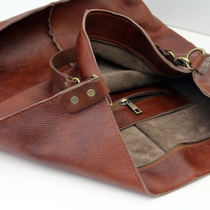 May include: A brown leather tote bag with a zipped pocket and a long strap. The bag is open and the inside is lined with a brown fabric.