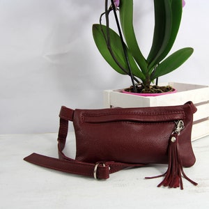 Puede incluir: Una ri&ntilde;onera de cuero burdeos con una hebilla plateada y un borla. La bolsa tiene un bolsillo con cremallera en la parte delantera.