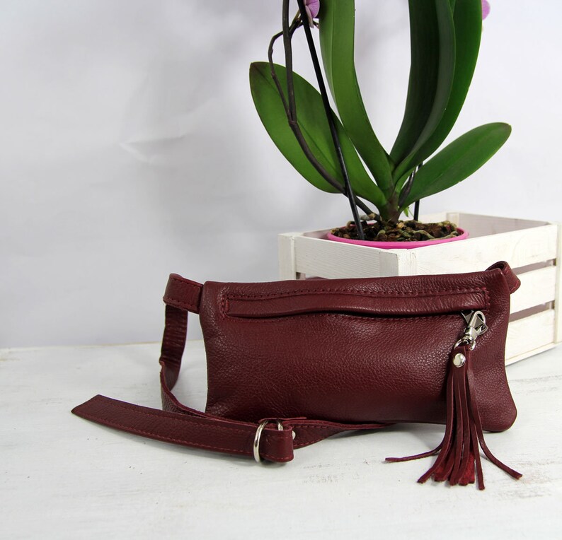 Puede incluir: Una ri&ntilde;onera de cuero burdeos con una hebilla plateada y un borla. La bolsa tiene un bolsillo con cremallera en la parte delantera.