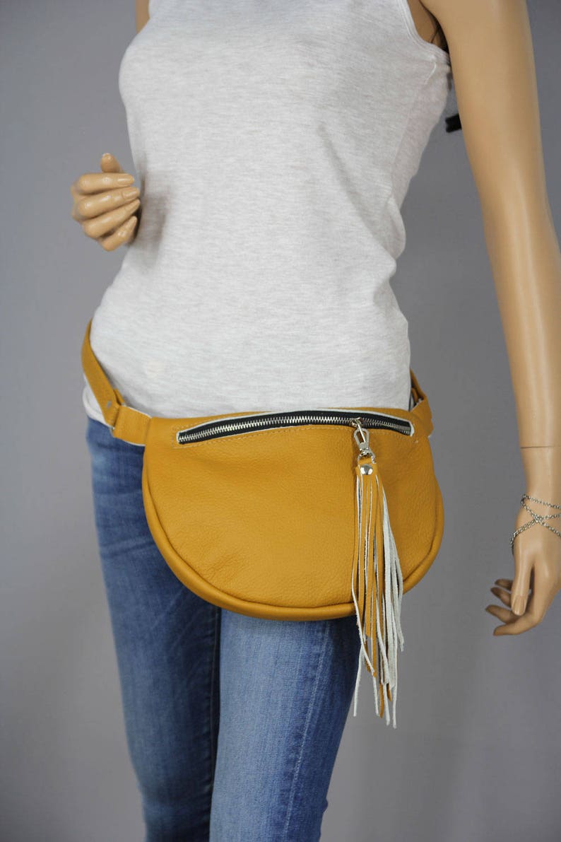 May include: A mustard yellow leather fanny pack with a white fringe tassel. The fanny pack has a black zipper and a silver zipper pull.