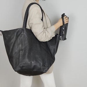 May include: A large black leather tote bag with a top handle and a zippered pocket on the side. The bag is being held by a person.
