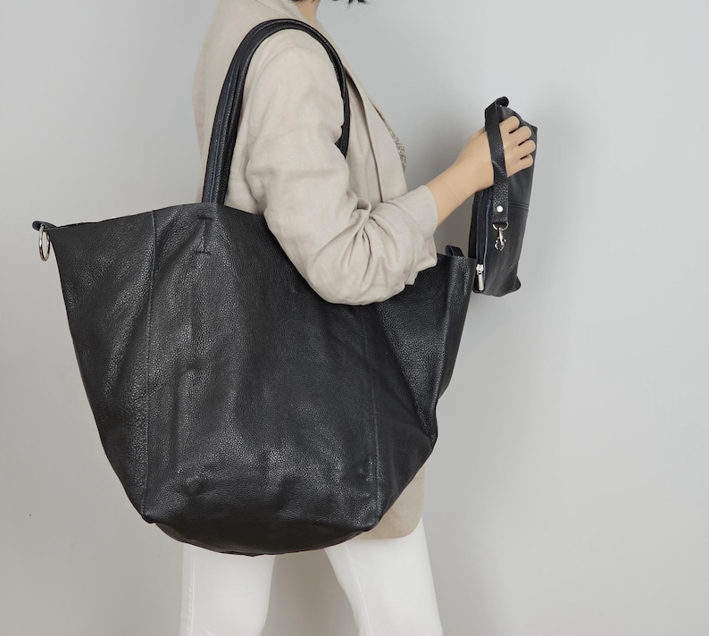 May include: A large black leather tote bag with a top handle and a zippered pocket on the side. The bag is being held by a person.