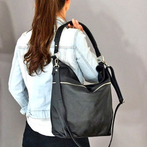 Op de afbeelding: Een zwarte leren tote bag met een lange schouderband. De tas heeft een ritsvak aan de voorkant en een klep sluiting.