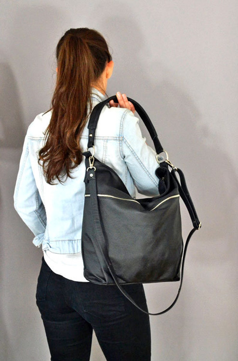 Op de afbeelding: Een zwarte leren tote bag met een lange schouderband. De tas heeft een ritsvak aan de voorkant en een klep sluiting.