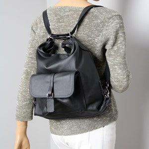 K&ouml;nnte beinhalten: Schwarzer Lederrucksack mit einer Klappentasche und verstellbaren Tr&auml;gern. Der Rucksack hat eine abgerundete Oberseite und silberfarbene Beschl&auml;ge. Die Tasche wird auf einer Schaufensterpuppe mit grauem Pullover und wei&szlig;er Hose gezeigt.