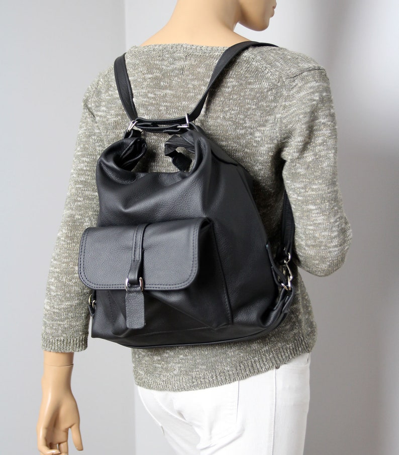 K&ouml;nnte beinhalten: Schwarzer Lederrucksack mit einer Klappentasche und verstellbaren Tr&auml;gern. Der Rucksack hat eine abgerundete Oberseite und silberfarbene Beschl&auml;ge. Die Tasche wird auf einer Schaufensterpuppe mit grauem Pullover und wei&szlig;er Hose gezeigt.
