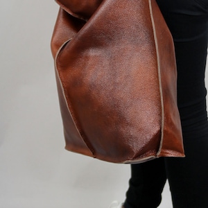 May include: A brown leather tote bag with a light brown trim. The bag is worn over the shoulder of a person wearing black trousers and white trainers.