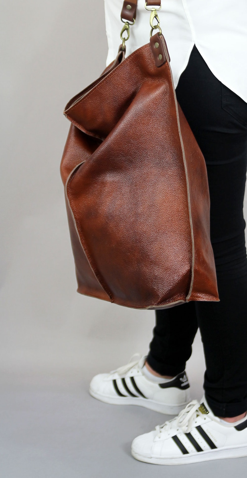 May include: A brown leather tote bag with a light brown trim. The bag is worn over the shoulder of a person wearing black trousers and white trainers.