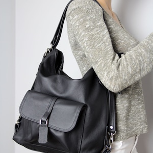 K&ouml;nnte beinhalten: Schwarze Leder-Umh&auml;ngetasche mit gro&szlig;er Fronttasche und Klappenverschluss. Die Tasche hat einen langen, verstellbaren Riemen und silberfarbene Hardware. Die Tasche wird auf einer Schaufensterpuppe gezeigt.