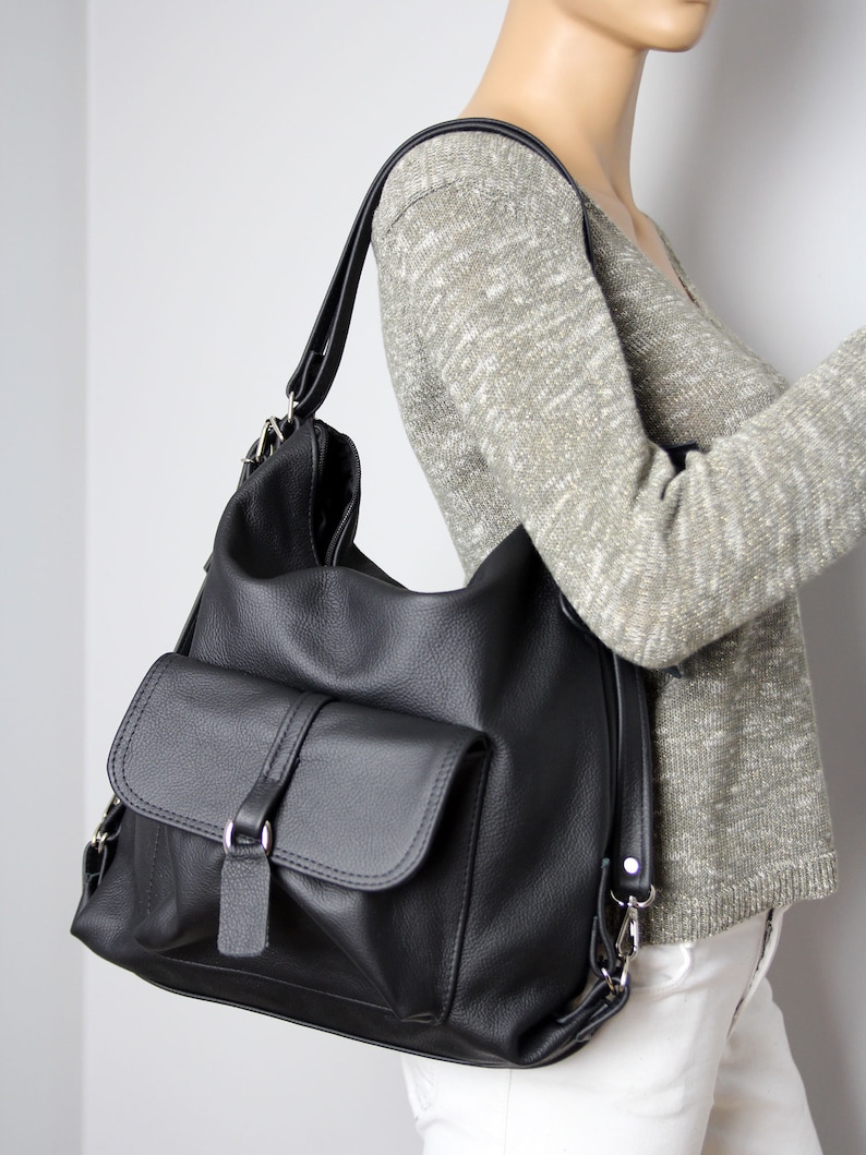 K&ouml;nnte beinhalten: Schwarze Leder-Umh&auml;ngetasche mit gro&szlig;er Fronttasche und Klappenverschluss. Die Tasche hat einen langen, verstellbaren Riemen und silberfarbene Hardware. Die Tasche wird auf einer Schaufensterpuppe gezeigt.
