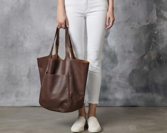 May include: A large, dark brown leather tote bag with two shoulder straps and a prominent exterior pocket on the front. The bag features a smooth, matte finish and a spacious, open-top design, providing a functional and simple aesthetic.