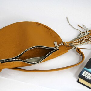 May include: A tan leather half-moon shaped crossbody bag with a zipper closure and a long fringe tassel.
