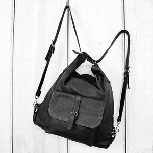 K&ouml;nnte beinhalten: Schwarzer Lederrucksack mit einer Frontklappentasche und verstellbaren Tr&auml;gern. Die Tasche h&auml;ngt vor einem wei&szlig;en Holzhintergrund. Der Rucksack hat ein l&auml;ssiges Design und einen Schnallenverschluss an der Vordertasche.