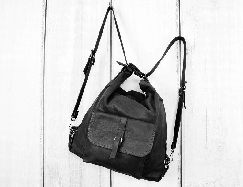 K&ouml;nnte beinhalten: Schwarzer Lederrucksack mit einer Frontklappentasche und verstellbaren Tr&auml;gern. Die Tasche h&auml;ngt vor einem wei&szlig;en Holzhintergrund. Der Rucksack hat ein l&auml;ssiges Design und einen Schnallenverschluss an der Vordertasche.