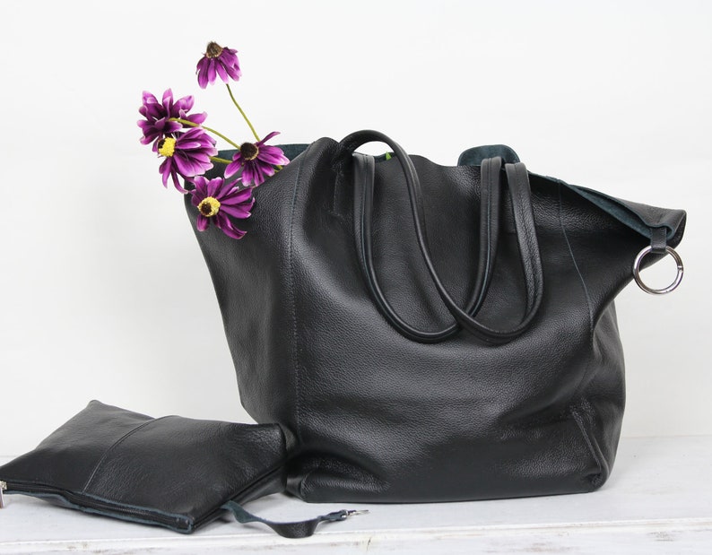 May include: A large black leather tote bag with two handles and a silver ring on the side. A small black leather pouch is in front of the bag. Purple flowers are sticking out of the top of the bag.
