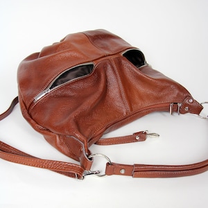 Pu&ograve; includere: Una borsa hobo in pelle marrone con una lunga tracolla regolabile. La borsa ha due tasche con cerniera sul davanti.
