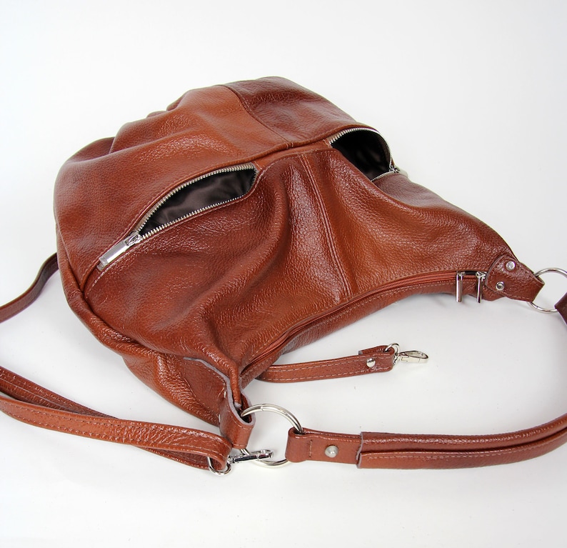 Pu&ograve; includere: Una borsa hobo in pelle marrone con una lunga tracolla regolabile. La borsa ha due tasche con cerniera sul davanti.