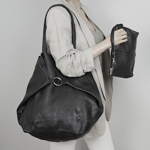 May include: A black leather tote bag with a silver ring detail. The bag has a slouchy, unstructured shape and is being held by a person.