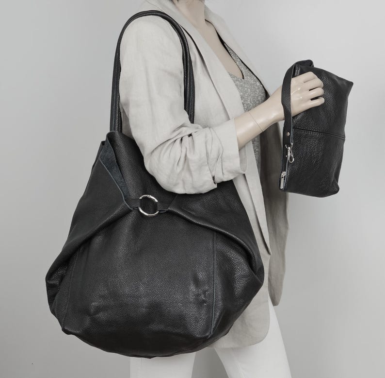 May include: A black leather tote bag with a silver ring detail. The bag has a slouchy, unstructured shape and is being held by a person.