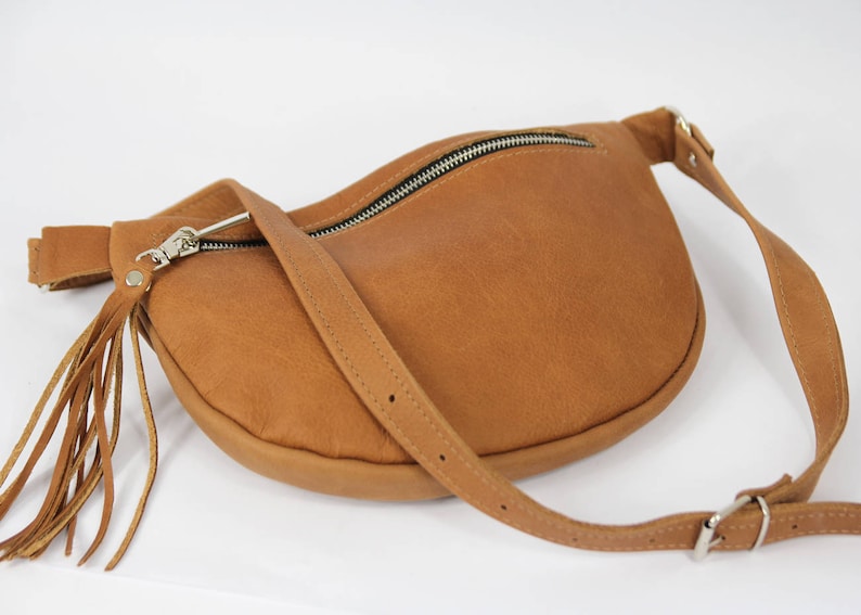 May include: A brown leather fanny pack with a zipper closure and a long fringe tassel. The bag has a long adjustable strap.