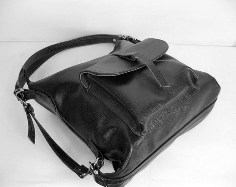 K&ouml;nnte beinhalten: Schwarzer Lederrucksack mit Klappenverschluss und verstellbaren Tr&auml;gern. Die Tasche hat eine strukturierte Oberfl&auml;che und einen einzelnen oberen Griff. Das Design umfasst silberfarbene Metallbeschl&auml;ge und eine Vordertasche unter der Klappe.