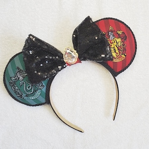 May include: A black sequined bow headband with two round ears featuring the Gryffindor and Slytherin house crests. The headband is tan and the ears are red and green with black trim. A small charm is centered on the bow.