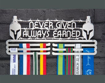 May include: A silver medal display rack with the words "Never Given Always Earned" and two Spartan helmet silhouettes. The rack has five rows for hanging medals and ribbons. The rack is mounted on a brick wall.